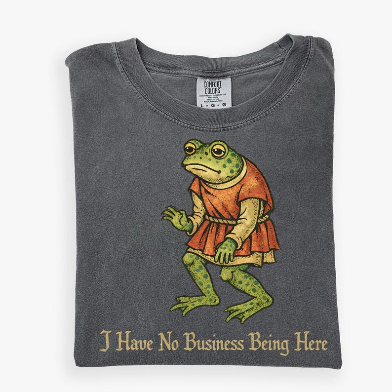 A t-shirt design showing a green frog standing upright in a medieval-style red tunic. Below the illustration is the phrase “I Have No Business Being Here” in black gothic-style lettering. The artwork has a vintage, illustrated appearance and is printed on a t-shirt.