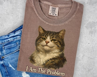 May include: A dusty rose-colored t-shirt featuring a smug-looking cat portrait with the text "I Am The Problem" in a serif font. The shirt is a Comfort Colors XL-TG-EG. The shirt is paired with blue denim shorts.