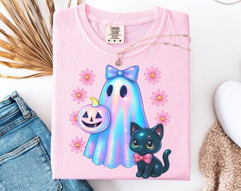 May include: A pale pink Comfort Colors t-shirt with a cartoon ghost wearing a blue bow, holding a jack-o'-lantern, and a black cat with a pink bow tie. The shirt is decorated with pink flowers and a gold necklace.