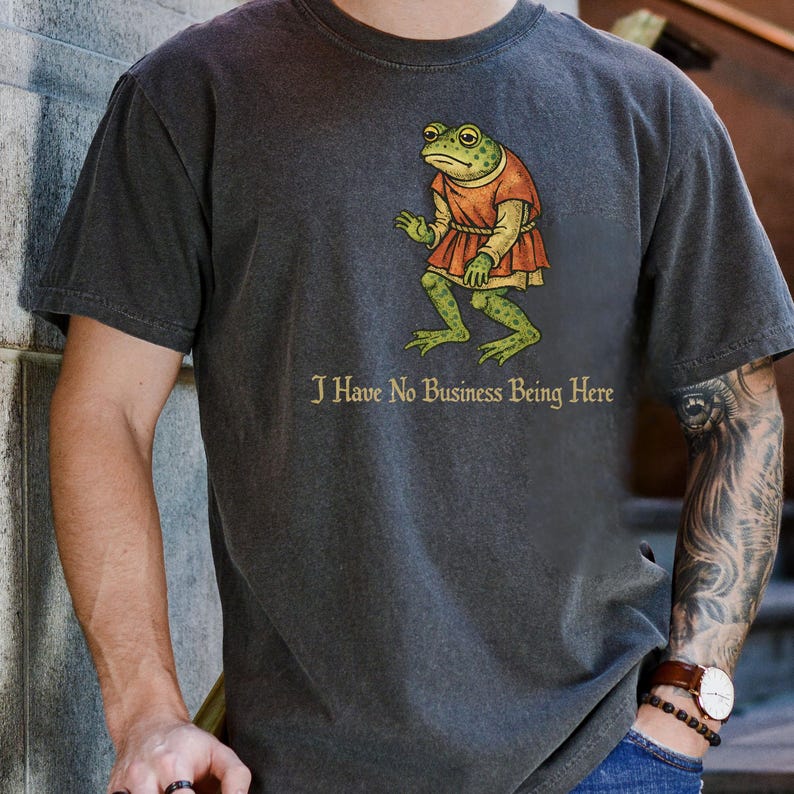 A t-shirt design showing a green frog standing upright in a medieval-style red tunic. Below the illustration is the phrase “I Have No Business Being Here” in black gothic-style lettering. The artwork has a vintage, illustrated appearance and is printed on a t-shirt.