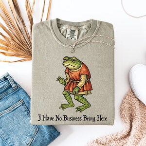 A t-shirt design showing a green frog standing upright in a medieval-style red tunic. Below the illustration is the phrase “I Have No Business Being Here” in black gothic-style lettering. The artwork has a vintage, illustrated appearance and is printed on a t-shirt.