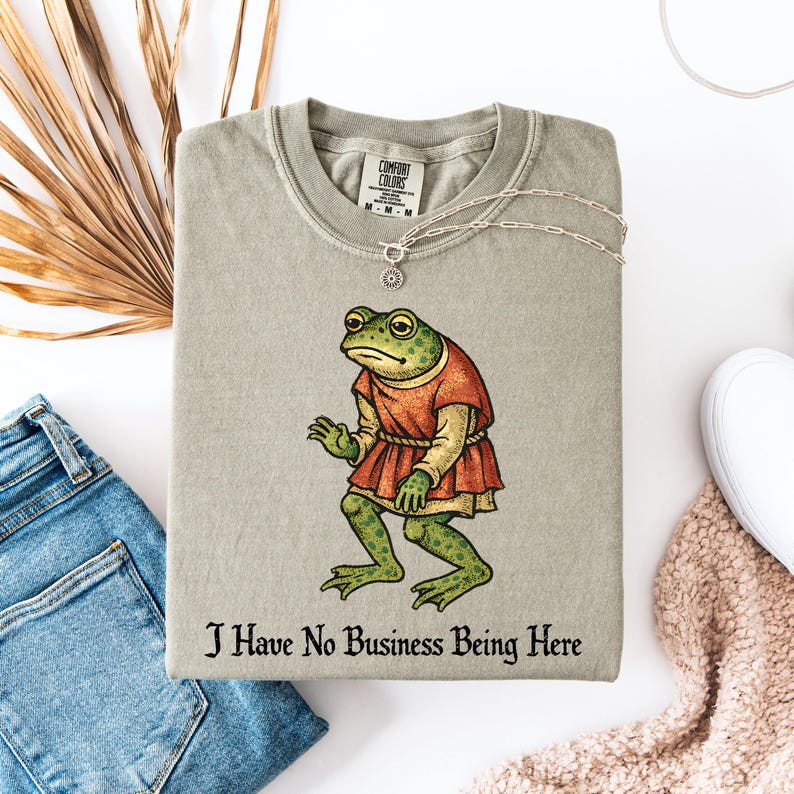 A t-shirt design showing a green frog standing upright in a medieval-style red tunic. Below the illustration is the phrase “I Have No Business Being Here” in black gothic-style lettering. The artwork has a vintage, illustrated appearance and is printed on a t-shirt.