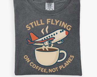 May include: Dark gray t-shirt with a graphic of an airplane taking off from a coffee cup. The text reads "STILL FLYING ON COFFEE, NOT PLANES" in a tan color. The shirt has a crew neck and short sleeves.