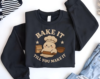 May include: Black sweatshirt with a graphic that says "Bake it Till You Make It". The design features a smiling dough character wearing a chef's hat, rolling dough with a rolling pin, with a flour bag and bowl.