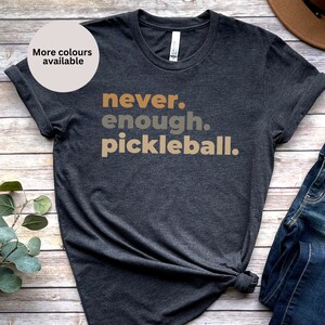 May include: A dark gray t-shirt with the text "never. enough. pickleball." printed in brown, orange, and beige.