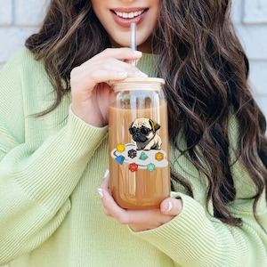 May include: A clear glass tumbler with a bamboo lid and a colorful sticker of a pug wearing sunglasses and sitting on a cloud of dice. The tumbler is filled with a brown liquid.