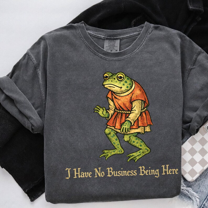 A t-shirt design showing a green frog standing upright in a medieval-style red tunic. Below the illustration is the phrase “I Have No Business Being Here” in black gothic-style lettering. The artwork has a vintage, illustrated appearance and is printed on a t-shirt.