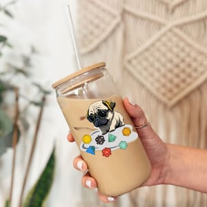 May include: A clear glass tumbler with a wooden lid and a straw. The tumbler is filled with a light brown beverage. A sticker of a pug wearing sunglasses and sitting on a cloud with dice is on the side of the tumbler.