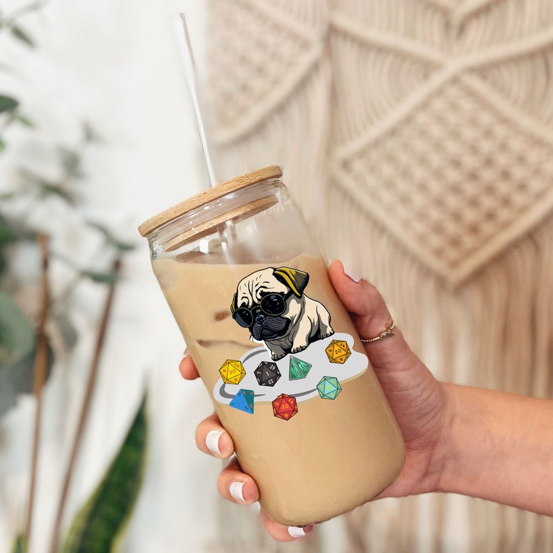 May include: A clear glass tumbler with a wooden lid and a straw. The tumbler is filled with a light brown beverage. A sticker of a pug wearing sunglasses and sitting on a cloud with dice is on the side of the tumbler.
