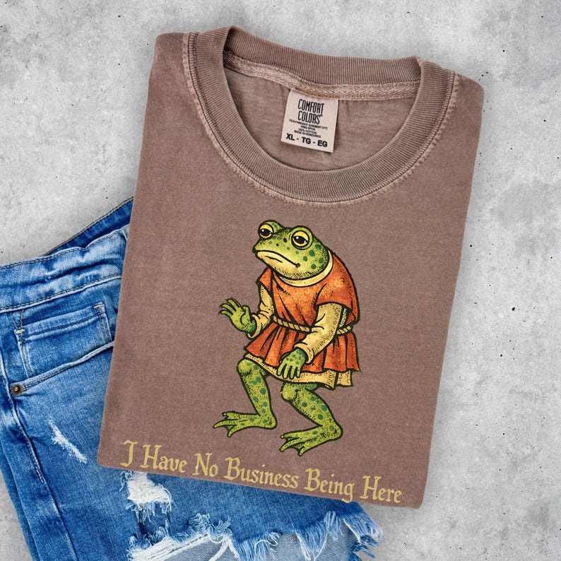 A t-shirt design showing a green frog standing upright in a medieval-style red tunic. Below the illustration is the phrase “I Have No Business Being Here” in black gothic-style lettering. The artwork has a vintage, illustrated appearance and is printed on a t-shirt.