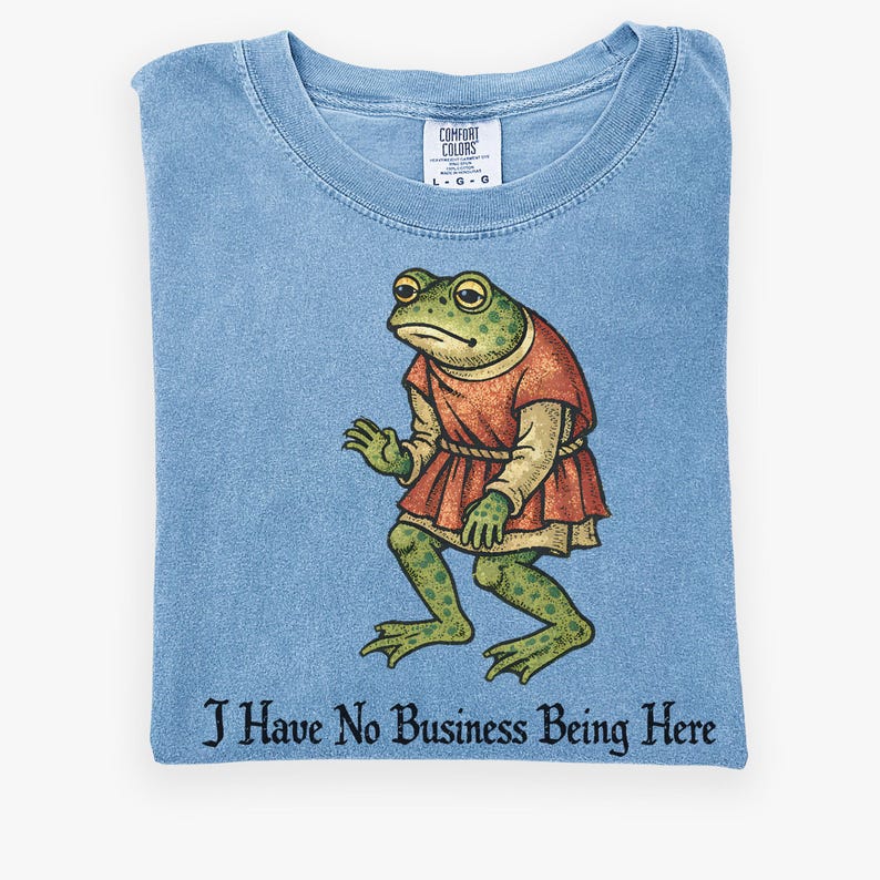 A t-shirt design showing a green frog standing upright in a medieval-style red tunic. Below the illustration is the phrase “I Have No Business Being Here” in black gothic-style lettering. The artwork has a vintage, illustrated appearance and is printed on a t-shirt.