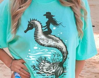 May include: A turquoise t-shirt featuring a seahorse design with a mermaid riding it. The seahorse has a detailed interior with a coral reef scene. The text "ISLAND REEF" is printed at the bottom of the shirt.