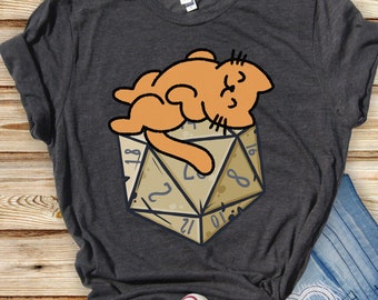 May include: A dark gray t-shirt with a cartoon illustration of an orange cat sleeping on top of a large, brown, twenty-sided die.