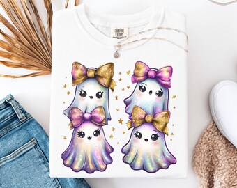 May include: White Comfort Colors t-shirt featuring four cartoon ghosts with bows and stars. The ghosts have a cute design with purple, gold, and iridescent colors. A silver necklace with a pendant is on the shirt.