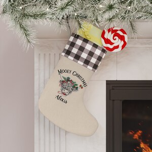 May include: A beige Christmas stocking with a black and white checked cuff. The stocking features a Highland cow graphic with the text "Mooney Christmas" and a name. A red and white swirled lollipop and a wrapped gift are visible.