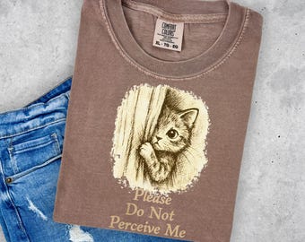 May include: A brown Comfort Colors t-shirt featuring a graphic of a kitten peeking from behind a curtain. The text "Please Do Not Perceive Me" is below the graphic. The shirt is paired with blue denim shorts.