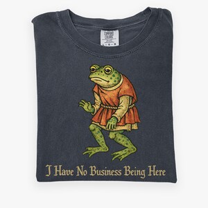 A t-shirt design showing a green frog standing upright in a medieval-style red tunic. Below the illustration is the phrase “I Have No Business Being Here” in black gothic-style lettering. The artwork has a vintage, illustrated appearance and is printed on a t-shirt.
