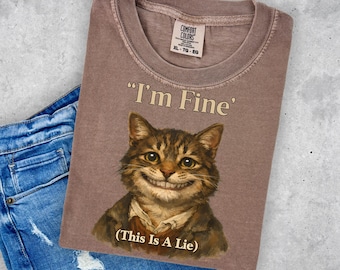 May include: A mauve-colored t-shirt with the text "I'm Fine" and a smiling cat graphic. Below the text is "(This Is A Lie)". The shirt is size XL-TG-EG.