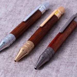 May include: Three wooden pens with silver, gold, and black metal accents. The pens have a dark wood finish and are arranged on a light grey fabric background.