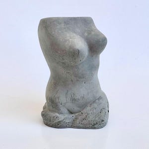 May include: A gray concrete sculpture of a female torso. The sculpture is smooth and has a rounded shape.