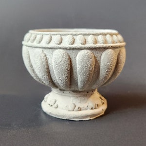 May include: A white ceramic planter with a decorative, fluted design. The planter has a round base and a wide, open top.