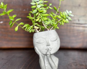 May include: A gray concrete planter shaped like a face with closed eyes and a finger to the lips, holding a green succulent plant. The text "Concrete Hephaestus" is at the bottom. The planter is on a dark wooden surface.