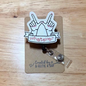 May include: A white badge reel with a design of two hands making a peace sign, with the word "whatever" on a banner. The badge reel is attached to a brown card with the text "Created by A FELTIE A DAY".