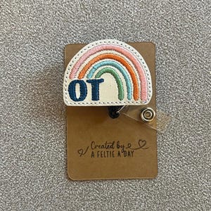 May include: A white rainbow with pink, orange, green, and blue embroidery. The rainbow has the letters "OT" embroidered in blue on the bottom. The rainbow is attached to a brown card with the text "Created by A Feltie A Day".