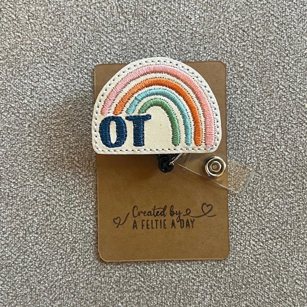May include: A white rainbow with pink, orange, green, and blue embroidery. The rainbow has the letters "OT" embroidered in blue on the bottom. The rainbow is attached to a brown card with the text "Created by A Feltie A Day".