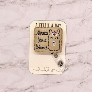 May include: A beige fabric badge holder with a gold embroidered alpaca wearing a nurse's hat. The text "Alpaca Your Wound" is embroidered on the badge holder. The text "A Feeltie A Day" is printed on the card behind the badge holder.