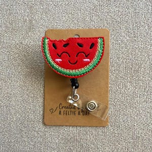 May include: A red watermelon slice badge reel with a smiling face and black seeds, outlined in green. The badge reel is attached to a brown card with the text "Created a feltie a day".