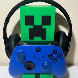 Puede incluir: Una figura de bloque verde con ojos y boca negros, sosteniendo un mando de Xbox azul. Los auriculares negros se colocan alrededor de la cabeza de la figura. La figura es un Creeper de Minecraft.