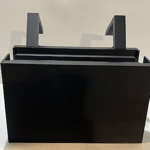 Könnte beinhalten: Ein schwarzes, rechteckiges, 3D-gedrucktes Objekt mit zwei grauen, L-förmigen Griffen. Das Objekt hat eine kastenartige Struktur mit einer flachen Oberseite und einer soliden Vorderseite. Die Griffe sind oben positioniert und erstrecken sich nach oben. Der Artikel ist wahrscheinlich ein Schreibtisch-Organizer oder eine Aufbewahrungslösung.