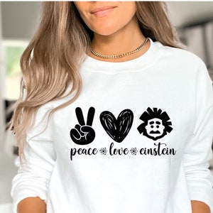 Puede incluir: Sudadera blanca con las palabras "peace love einstein" en letras negras y gráficos. Los gráficos incluyen un signo de la paz, un corazón y una imagen estilizada de Albert Einstein. La sudadera es de manga larga.