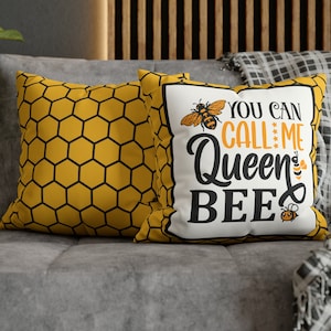 May include: Two decorative cushions on a grey sofa. One cushion features a honeycomb pattern in yellow and black. The other cushion has the text "YOU CAN CALL ME Queen BEE" with bee graphics in black, yellow, and orange.