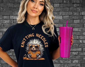 Op de afbeelding: Zwart t-shirt met een afbeelding van een egel met een zonnebril en een trui, met de tekst "Knitting Needles" in een cirkel. Een roze beker is ook zichtbaar.