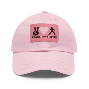 Op de afbeelding: Lichtroze baseballpet met een gebogen klep. De pet heeft een rechthoekige patch met een vredesteken, een hart en een Elvis-silhouet, met de woorden "PEACE LOVE ELVIS" in zwarte tekst.