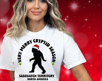 May include: A white t-shirt featuring a black silhouette of a Sasquatch wearing a red and white Santa hat. The text reads VERY MERRY CRYPTID SEASON, SASQUATCH TERRITORY, NORTH AMERICA, with small green and red holly leaf accents on the sides.