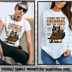 Op de afbeelding: Wit T-shirt en longsleeve shirt met een afbeelding van een wasbeer met een zonnebril die in een strandstoel zit. De tekst luidt: "I CAME FOR THE FIREWORKS, NOT THE PEOPLE." Het ontwerp bevat vuurwerk en patriottische kleuren.