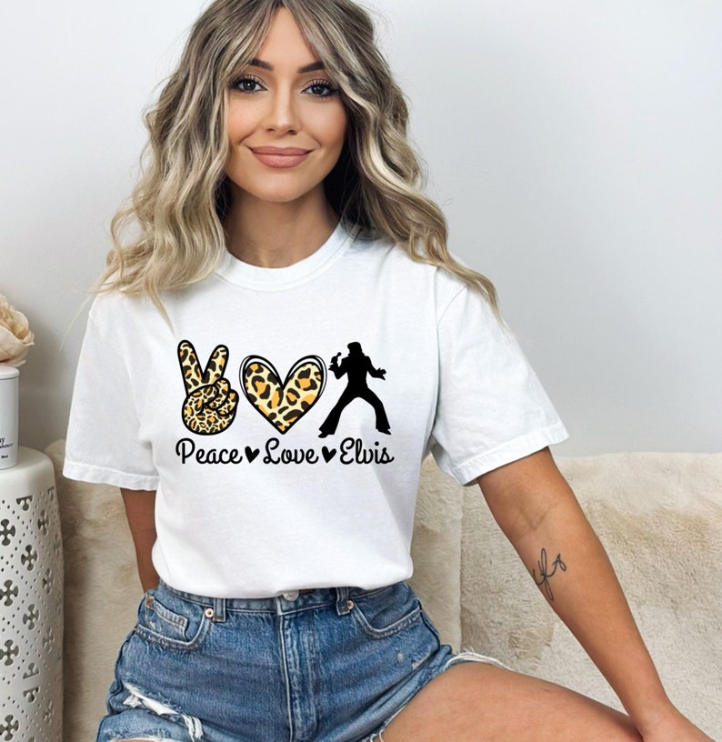 K&ouml;nnte beinhalten: Wei&szlig;es T-Shirt mit einem Peace-Zeichen, einem Herzen und einer Elvis-Silhouette. Das Peace-Zeichen und das Herz sind mit einem Leopardenmuster verziert. Der Text "Peace Love Elvis" ist unter dem Design gedruckt.