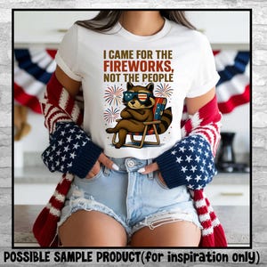 Op de afbeelding: Wit T-shirt met de tekst "I CAME FOR THE FIREWORKS, NOT THE PEOPLE." Het shirt heeft een cartoon wasbeer met een zonnebril die in een strandstoel zit. De wasbeer is bruin met een gestreepte staart. Vuurwerk staat op de achtergrond.
