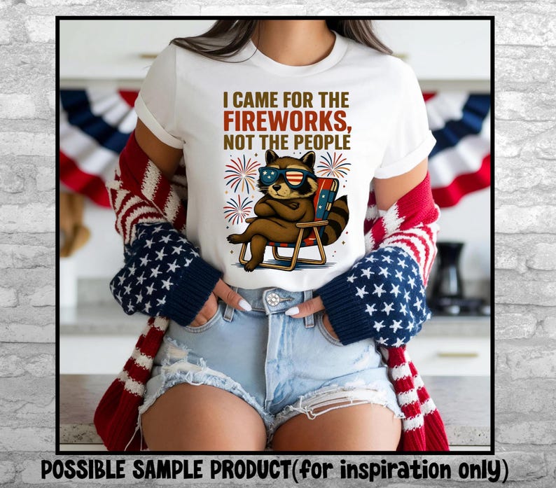 Op de afbeelding: Wit T-shirt met de tekst "I CAME FOR THE FIREWORKS, NOT THE PEOPLE." Het shirt heeft een cartoon wasbeer met een zonnebril die in een strandstoel zit. De wasbeer is bruin met een gestreepte staart. Vuurwerk staat op de achtergrond.