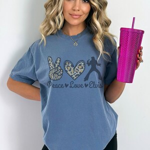 K&ouml;nnte beinhalten: Ein blaues T-Shirt mit einem Peace-Zeichen, einem Herzen und einer Elvis-Silhouette. Die Grafik ist in Schwarz, Braun und Wei&szlig; gedruckt. Der Text "Peace Love Elvis" ist unter der Grafik gedruckt.