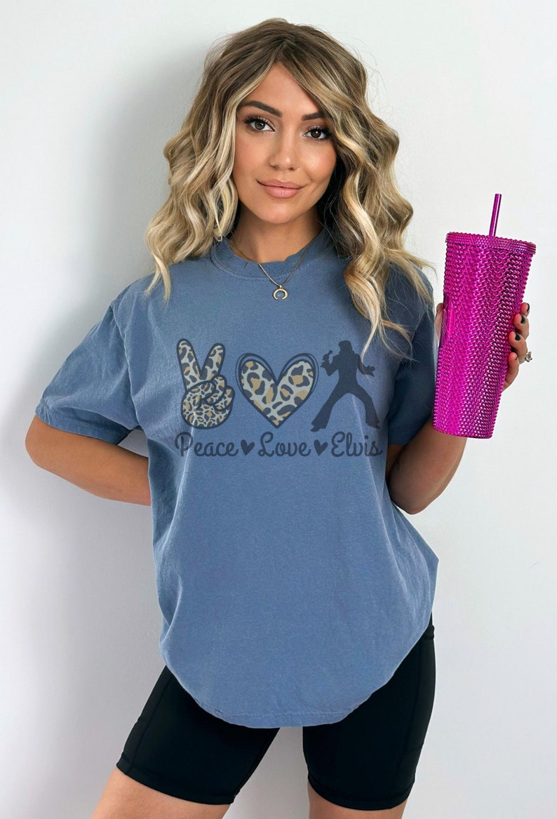 K&ouml;nnte beinhalten: Ein blaues T-Shirt mit einem Peace-Zeichen, einem Herzen und einer Elvis-Silhouette. Die Grafik ist in Schwarz, Braun und Wei&szlig; gedruckt. Der Text "Peace Love Elvis" ist unter der Grafik gedruckt.
