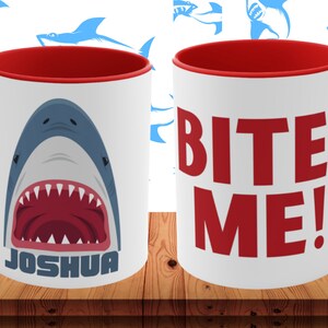 Könnte beinhalten: Weiße Keramik-Tasse mit rotem Innenraum und Griff. Eine Seite zeigt einen Cartoon-Hai mit offenem Maul und dem Namen "JOSHUA". Die andere Seite hat die Worte "BITE ME!" in Rot. Der Hintergrund hat ein Muster aus blauen Haien.