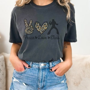 K&ouml;nnte beinhalten: Ein dunkelgraues T-Shirt mit einem Peace-Zeichen, einem Herzen und einer Silhouette von Elvis Presley. Das Peace-Zeichen und das Herz sind mit einem Leopardenmuster verziert. Der Text "Peace Love Elvis" ist unter der Grafik gedruckt.