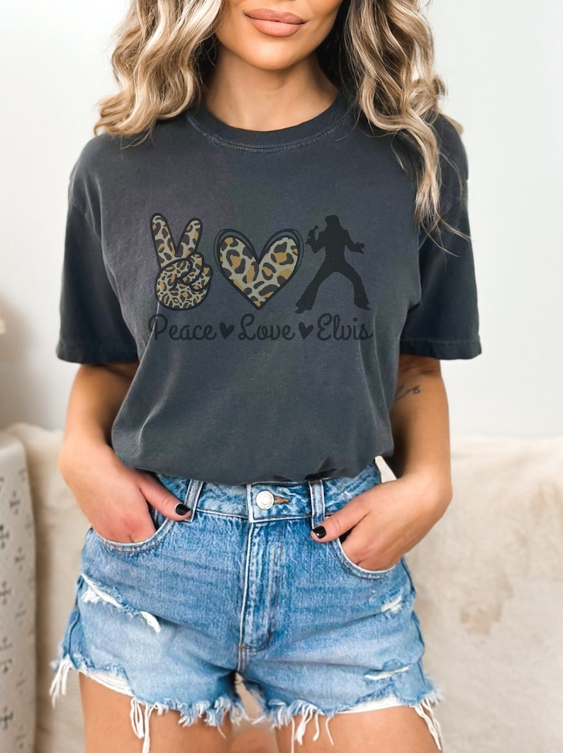K&ouml;nnte beinhalten: Ein dunkelgraues T-Shirt mit einem Peace-Zeichen, einem Herzen und einer Silhouette von Elvis Presley. Das Peace-Zeichen und das Herz sind mit einem Leopardenmuster verziert. Der Text "Peace Love Elvis" ist unter der Grafik gedruckt.