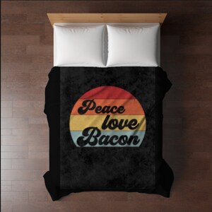 May include: A black comforter with a retro design featuring a red, orange, yellow, and blue striped circle. The words "Peace love Bacon" are written in a cursive font within the circle. Two white pillows are visible.
