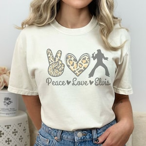 K&ouml;nnte beinhalten: Ein wei&szlig;es T-Shirt mit einem Peace-Zeichen, einem Herzen und einer Elvis-Silhouette. Das Peace-Zeichen und das Herz sind mit einem Leopardenmuster bedruckt. Der Text "Peace Love Elvis" ist unter dem Motiv gedruckt.