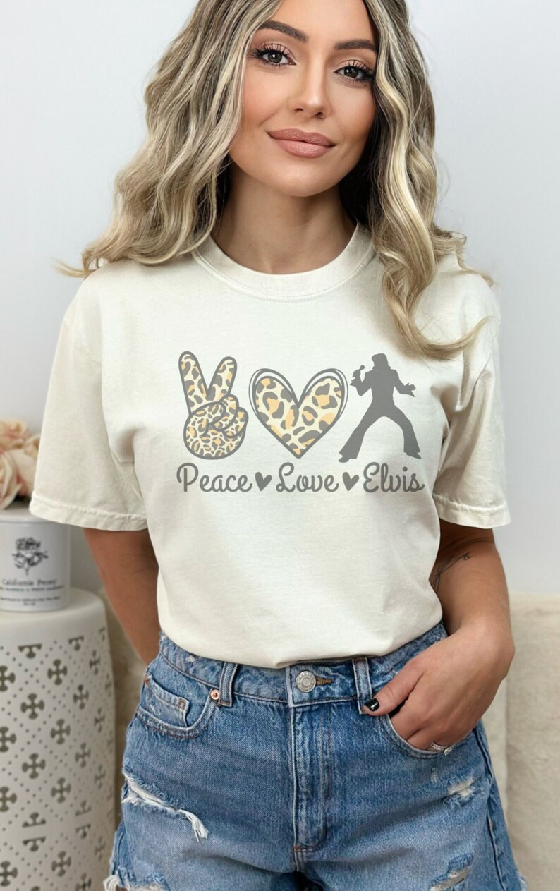 K&ouml;nnte beinhalten: Ein wei&szlig;es T-Shirt mit einem Peace-Zeichen, einem Herzen und einer Elvis-Silhouette. Das Peace-Zeichen und das Herz sind mit einem Leopardenmuster bedruckt. Der Text "Peace Love Elvis" ist unter dem Motiv gedruckt.